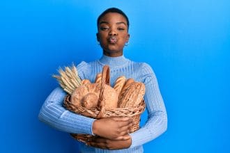 Young african american woman holding wicker basket with bread puffing cheeks with funny face. mouth inflated with air, catching air.
